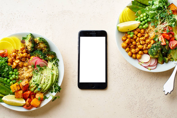 Flat lay with black smartphone and healthy vegetarian lunch bowls with avocado and chickpeas with quinoa and vegetables garnished with microgreens and nut dressing. Nutrient food delivery concept  - Zdjęcie, obraz