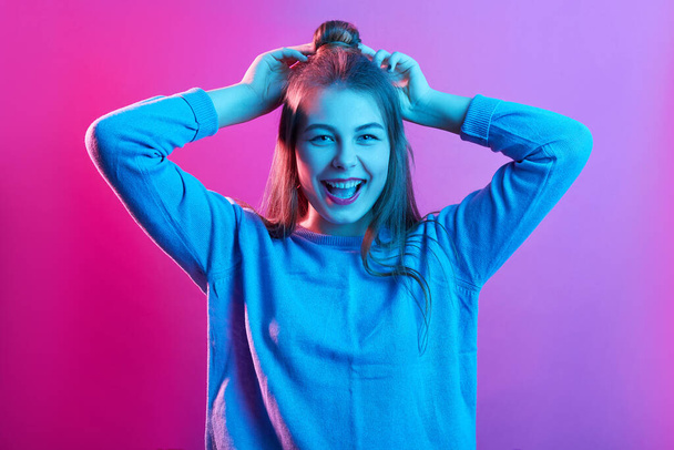 Horizontal shot of happy laughing teenager girl standing with hands up, has hair bun, wearing casual sweater,posing isolated over pink neon background. People sincere emotions, lifestyle concept. - Фото, зображення