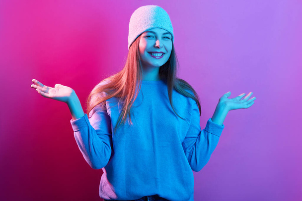 Closeup portrait of happy woman spreading hands aside, dresses casual jumper and headdress, looks happy and satisfied, posing isolated on pink neon background, looks smiling at camera. People emotions - Foto, Imagen