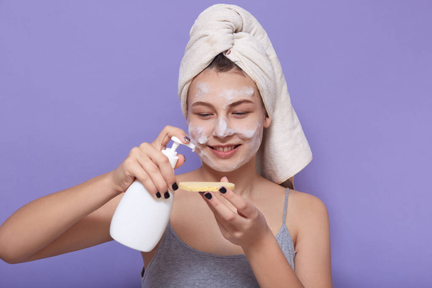 Indoor shot of young charming woman wrapped white towel using cleansing foam from dispenser, applies agent on cosmetic sponge, doing morning procedures, posing isolated over lilac background. - Foto, Imagem