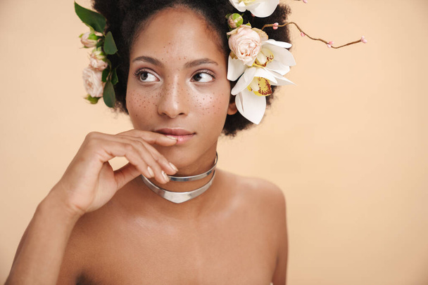 Portrait of young half-naked freckled african american woman with flowers in her hair isolated over beige background - Fotó, kép
