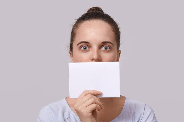 Studio shot of shocked woman with hair bun, holding empty card near her lips, posing with big eyes, covers her mouth with piece of paper with copy space for inscription, isolated on studio background. - Фото, изображение