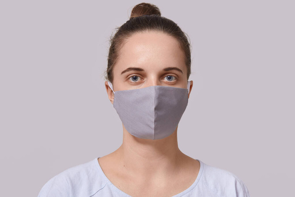 Closeup portrait of serious woman wearing gray protective mask, lady dresses white casual t shirt looking directly at camera, posing isolated over white background. Corona virus protection concept. - Foto, Bild