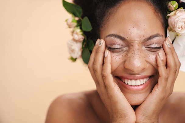 Portrait of young half-naked freckled african american woman with flowers in her hair isolated over beige background - Photo, image