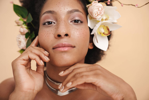 Portrait of young half-naked freckled african american woman with flowers in her hair isolated over beige background - Foto, Bild