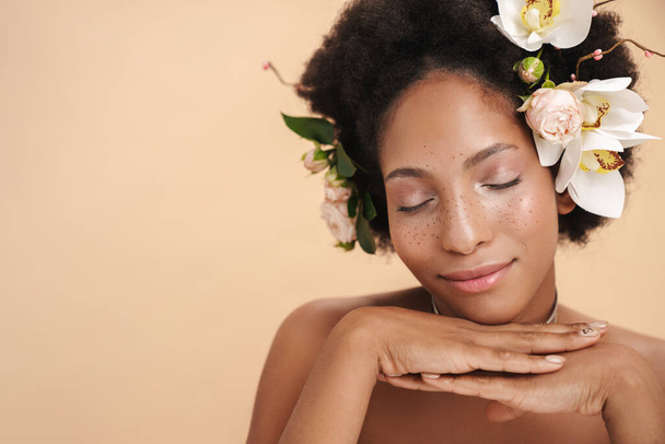 Portrait of young half-naked freckled african american woman with flowers in her hair isolated over beige background - Fotografie, Obrázek