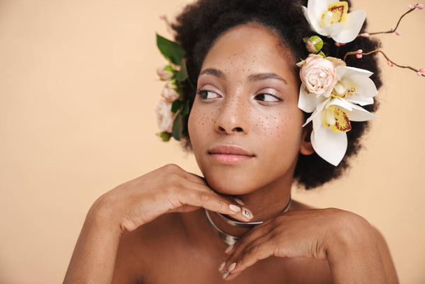 Portrait of young half-naked freckled african american woman with flowers in her hair isolated over beige background - Foto, imagen