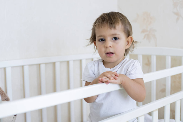 concept stay home quarantined, take care of your family. little cute toddler is standing in the crib at home and looking at the camera. place for copy space - Foto, Imagen