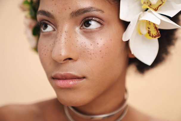 Portrait of young half-naked freckled african american woman with flowers in her hair isolated over beige background - Zdjęcie, obraz