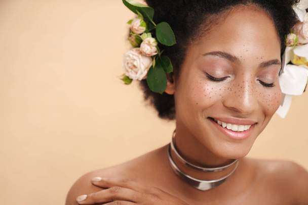 Portrait of young half-naked freckled african american woman with flowers in her hair isolated over beige background - 写真・画像
