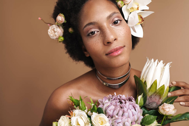 Portrait of young half-naked freckled african american woman posing with flowers isolated over beige background - Фото, зображення