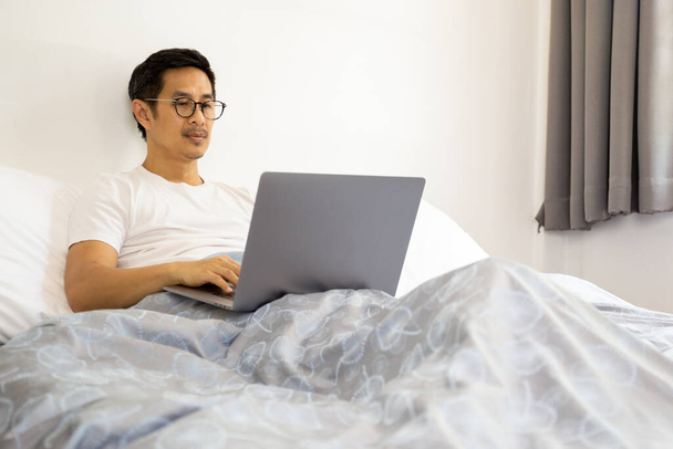 Knappe man met bril aan het werk op laptop in bed thuis covid-19 uitbraak - Foto, afbeelding