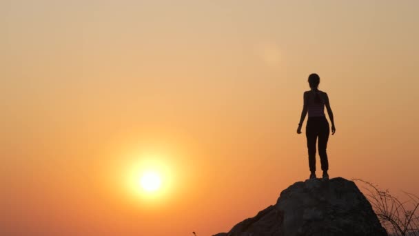 Silhouette d'une randonneuse sautant seule sur une grosse pierre au coucher du soleil dans les montagnes. Touriste femelle levant les mains sur le rocher élevé dans la nature du soir. Tourisme, voyage et mode de vie sain concept. - Séquence, vidéo