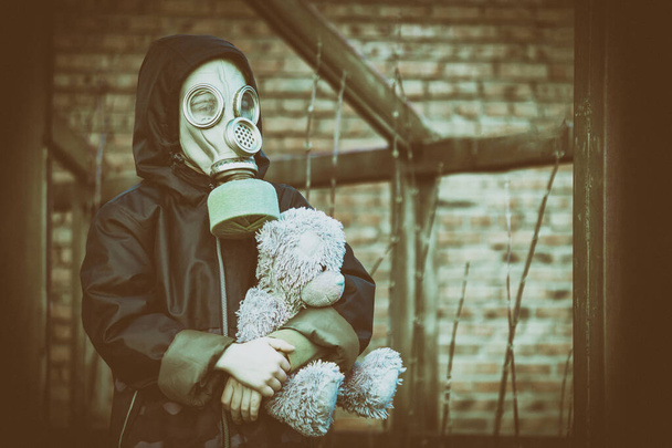 A small boy with a gas mask on his face and a hood on his head hugs his toy bear, against the ruins. Tinted photo with noise overlay, stylized as an old photo. The concept of air pollution, ecology. - Фото, изображение