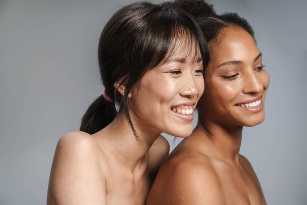 Portrait of two multinational half-naked women hugging and laughing isolated over grey background - Foto, imagen