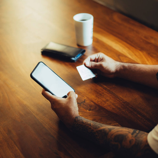 Mockup záběr na mužskou ruku s tetováním držícím mobilní telefon a kreditní kartu s prázdnou obrazovkou na stole v domácí kanceláři. Pro nákupy. - Fotografie, Obrázek