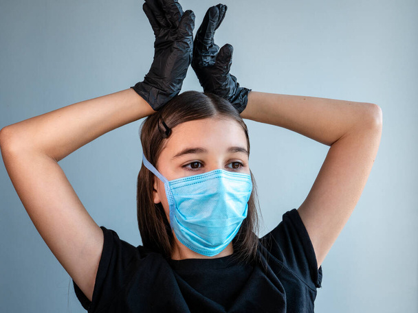 青い保護医療面マスクの女性の十代の少女は、彼女の頭の上に黒いラテックス手袋で彼女の手を保持することによってコロナを示しています。コロナウイルス｜Covid-19流行病の概念. - 写真・画像