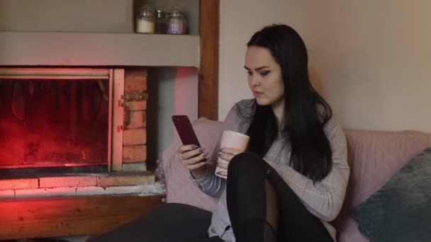 The brunette is sitting at home and looking at the phone, the girl is chatting, playing games, smiling. The girl sits dma near the fireplace, all the time on the phone. - Кадры, видео