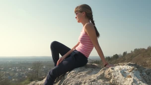 Młoda, zrelaksowana kobieta siedząca na świeżym powietrzu na wielkim kamieniu ciesząca się ciepłym letnim dniem. Dziewczyna medytująca i relaksująca się na łonie natury o zachodzie słońca. - Materiał filmowy, wideo