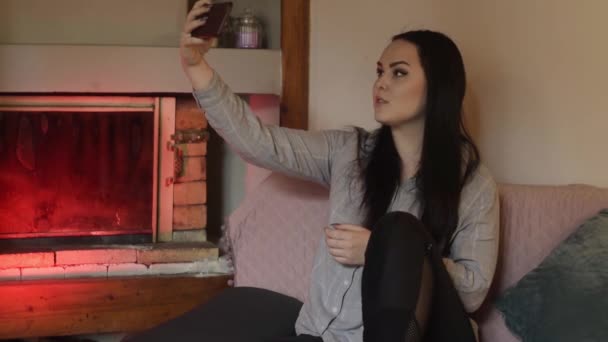Girl sitting at home on the sofa against the background of the fireplace. A girl takes a photo on the phone as she sits at home. - Footage, Video
