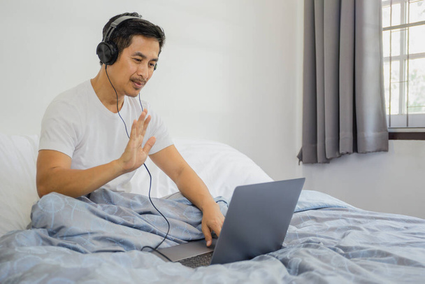 ヘッドフォンを持つ男がベッドに座って手を振ってビデオ通話を持っているラップトップのコロナウイルス検疫 - 写真・画像