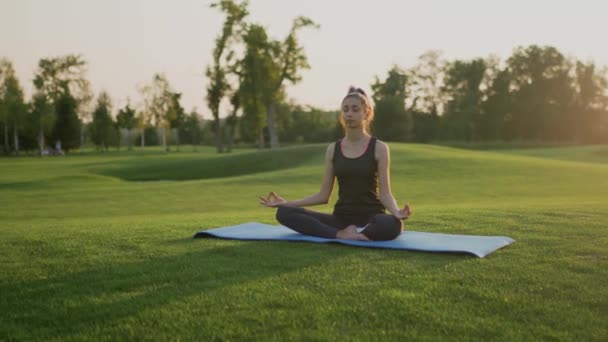 Junges Mädchen sitzt auf grünem Gras in Lotusposition, Yoga-Kurs bei Sonnenuntergang. - Filmmaterial, Video