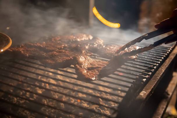夜は火の上でおいしいジューシーなステーキを焼くバーベキューグリル、手はトングを保持し、肉を裏返します。クローズアップ、ソフトフォーカス。煙が強調され - 写真・画像