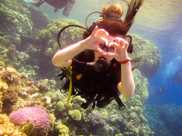 Lovely girl diver shows with a hand gesture a sign of love, a heart from fingers with a partner instructor for safe swimming under water in the Red Sea with coral reefs Egypt, Sharm El Sheikh. - Photo, Image
