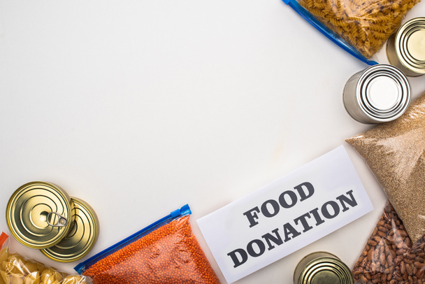 vista superior de latas e grumos em sacos de zíper perto do cartão com letras de doação de alimentos no fundo branco
 - Foto, Imagem