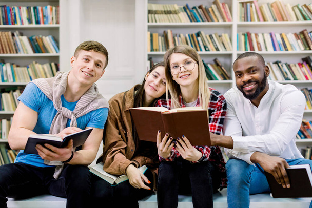 Vier multiethnische, gut gelaunte Studenten treffen sich in der Bibliothek, lesen Bücher, kommunizieren und verbringen Zeit miteinander. Glücklicher internationaler Büchertag - Foto, Bild