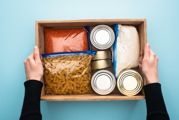 cropped view of woman holding wooden box with cans and groats in zipper bags on blue background - Photo, Image