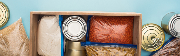 top view of cans and groats in zipper bags in wooden box on blue background, food donation concept - Φωτογραφία, εικόνα