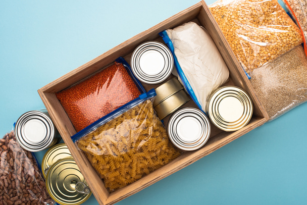 top view of cans and groats in zipper bags in wooden box on blue background, food donation concept - Photo, Image