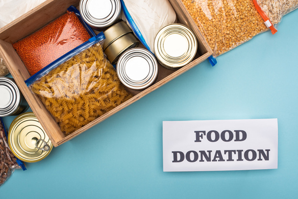 top view of cans and groats in zipper bags in wooden box near card with food donation lettering on blue background - Photo, Image