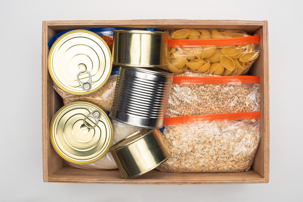 top view of cans and groats in zipper bags in wooden box on white background, food donation concept - Foto, afbeelding
