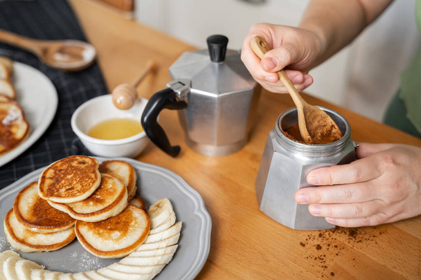 Hände einer jungen Frau, die in der Küche neben den Pfannkuchen gemahlenen Kaffee in eine Geysir-Kaffeemaschine stampft. Frühstück zu Hause kochen. - Foto, Bild