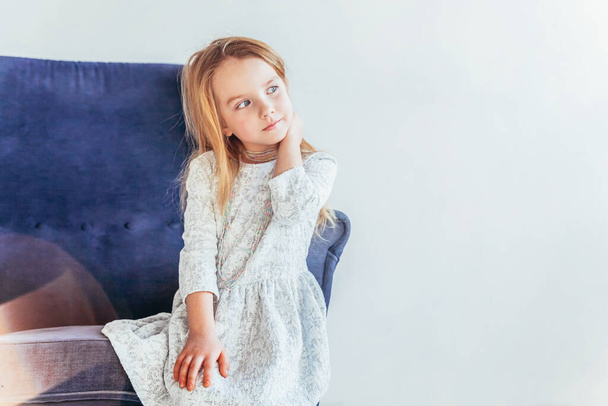 Bleiben Sie zu Hause, bleiben Sie sicher. Süßes kleines Mädchen in weißem Kleid sitzt auf einem modernen gemütlichen blauen Stuhl und entspannt es sich im weißen, hellen Wohnzimmer zu Hause drinnen. Kindheit Schüler Jugend entspannen Konzept - Foto, Bild