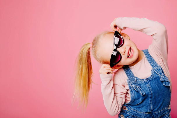 Young cute smilling blonde girl is holding her retro sunglasses on pink background. Concept of happy childhood. - Photo, Image