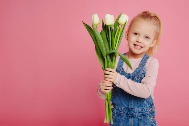 Young cute smiling girl is holding white tulips on pink background. Concept of happy childhood. Happy mother`s day or children`s day. - Photo, Image
