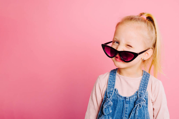 Jovem menina loira bonito smilling em óculos de sol retro está piscando no fundo rosa. Conceito de infância feliz
. - Foto, Imagem