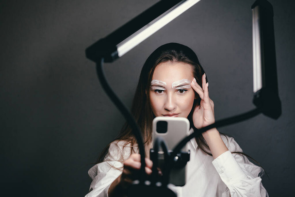 Beauty blogger girl quarantined at home doing eyebrow shaping with wax. Beauty lamp and a phone on a tripod - Foto, Imagem