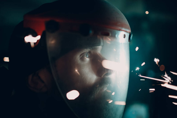 Homem barbudo adulto em máscara protetora transparente e moedor de serra com partículas de metal voadoras faíscas na escuridão
 - Foto, Imagem