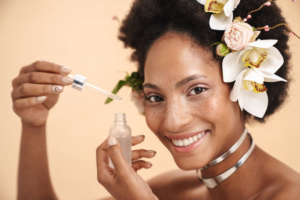 Portrait of young freckled african american woman holding facial serum cream bottle isolated over beige background - 写真・画像