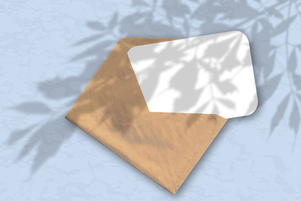 Una busta con un foglio di carta bianca strutturata sullo sfondo blu della parete. Il Mockup si sovrappone alle ombre delle piante. La luce naturale getta ombre dal fogliame dell'albero
. - Foto, immagini