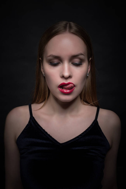 Fashionable young, seductive, emotional, beautiful girl, a woman with blond straight hair, red lipstick on her lips, licking her tongue, and a black dress on a black and dark background. - Foto, Imagen