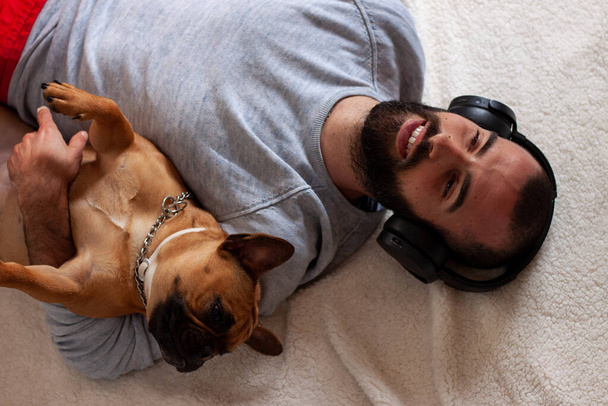 Французький бульдог з молодим чоловіком у навушниках посміхається, лежачи вдома на підлозі. - Фото, зображення