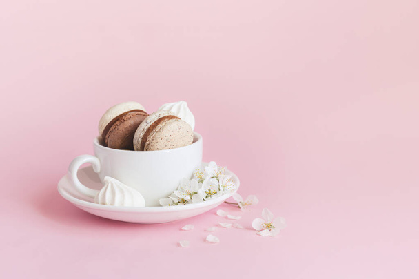 Beker met franse macarons, marshmallows en lentebloesem op een roze pastelachtergrond. Romantis lente achtergrond. - Foto, afbeelding