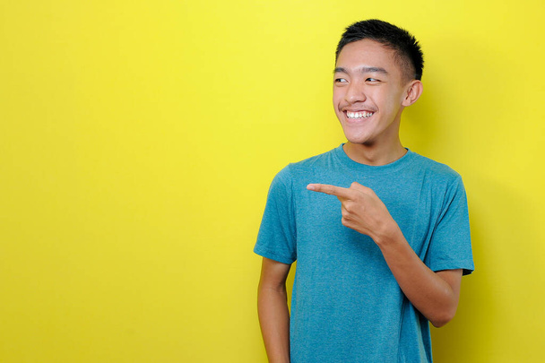 Happy Young Asian man in casual t-shirt wijzend op een kopieerruimte, geïsoleerd op gele achtergrond - Foto, afbeelding