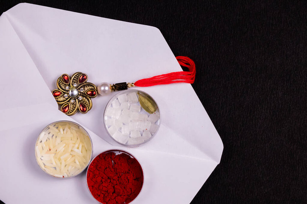 Концепция Индийского фестиваля - Элегантный Ракхи в белом конверте с рисовыми зернами, кумкумом, сахарной породой и кардамоном плоская жестяная коробка на черном фоне
 - Фото, изображение