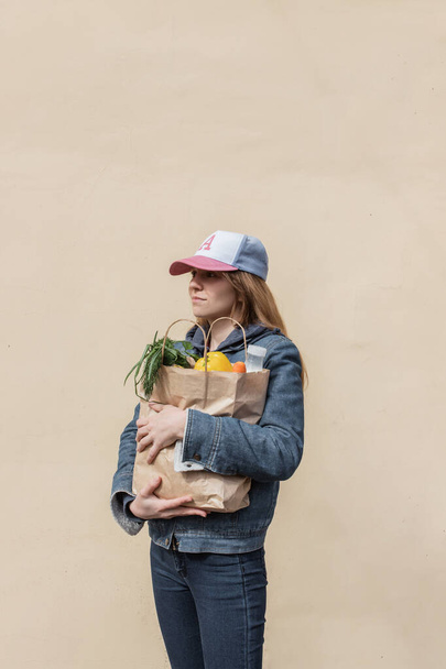 Smiling girl in cap and denim clothes isolated on yellow background with fresh vegetables and fruit in hands. Delivery service from shop or restaurant concept. Covid 19. Young woman grocery courier. - Foto, Imagen